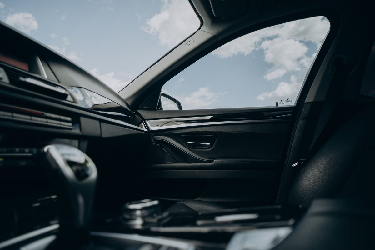 View from inside a car toward the driver's door: the side window opening with sky and clouds beyond, the door panel and its inside handle