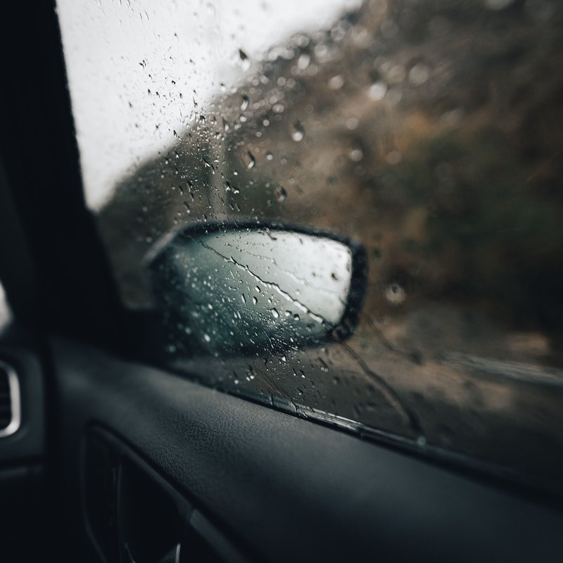 Raindrops on a car's front side window, the wet side mirror and roadside trees blurred beyond it