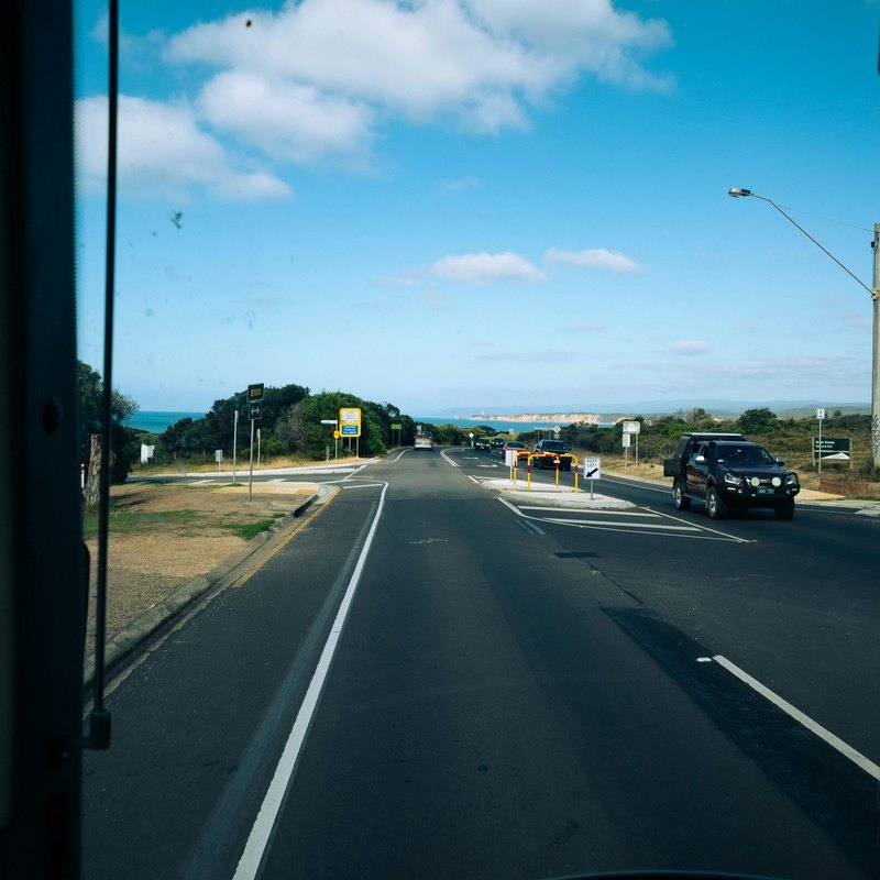 A two-lane road under a blue sky with white clouds, seen through a large windshield from the driver's seat