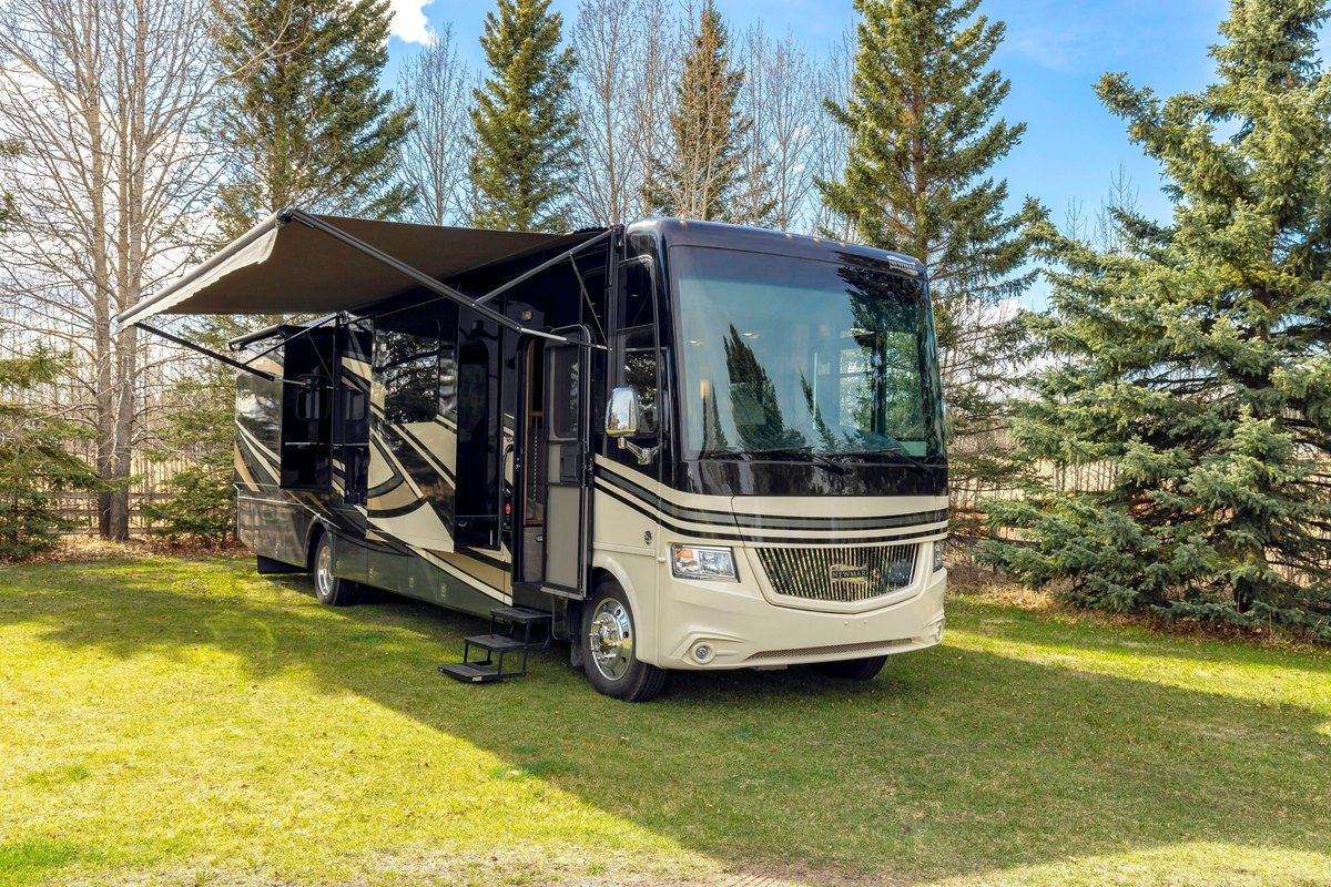 Tan and black Class A motorhome on a lawn with its awning out and a large one-piece windshield