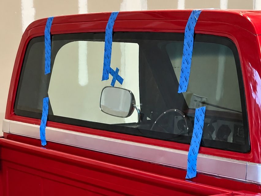 Close view from behind the cab of a red classic pickup, its newly fitted back glass held by blue tape strips