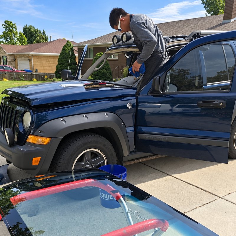 Young man wiping the empty windshield frame of a navy Jeep Liberty in a driveway
